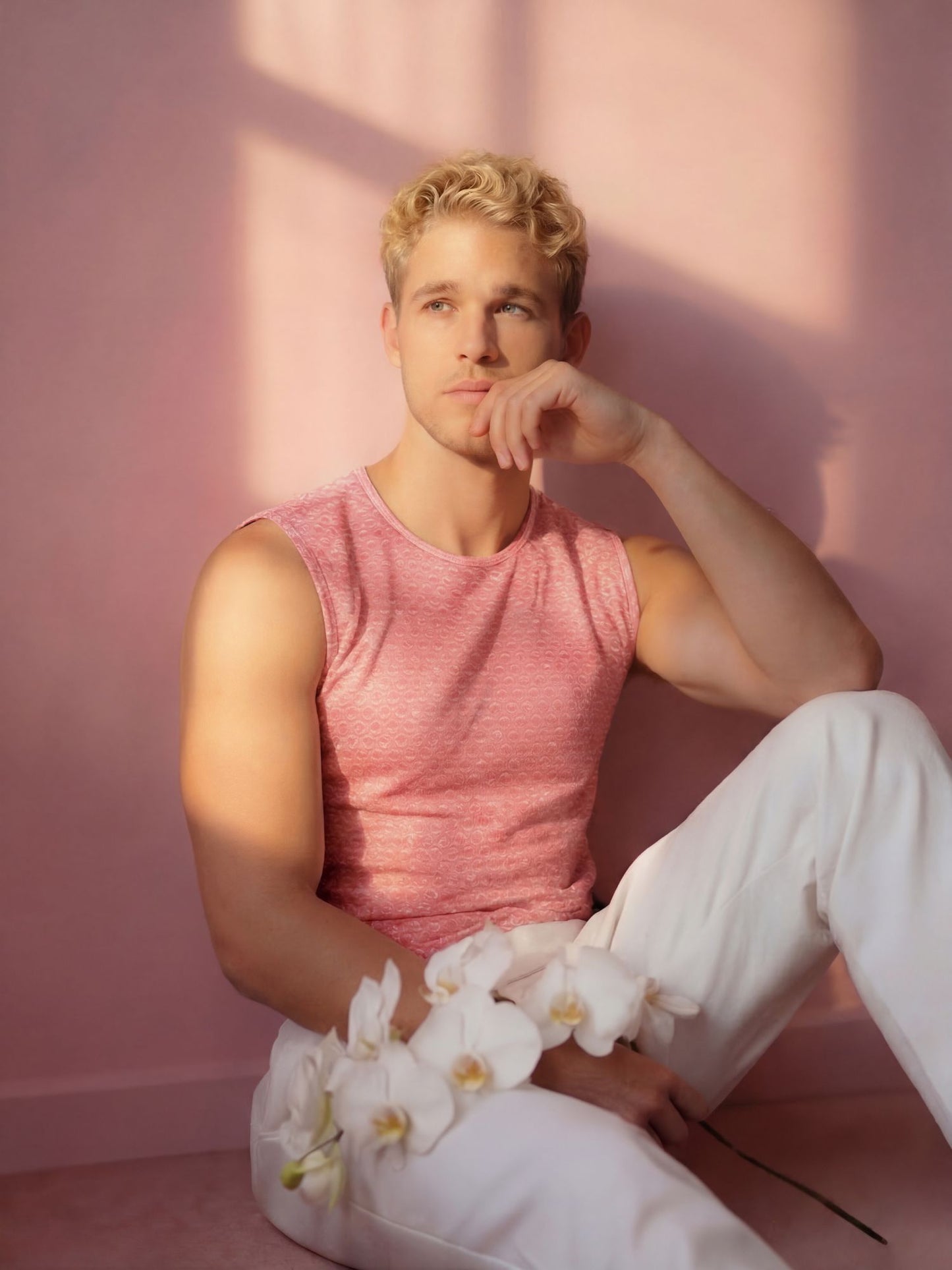 Man in pink tank top and white pants sitting against a pink wall with white flowers.