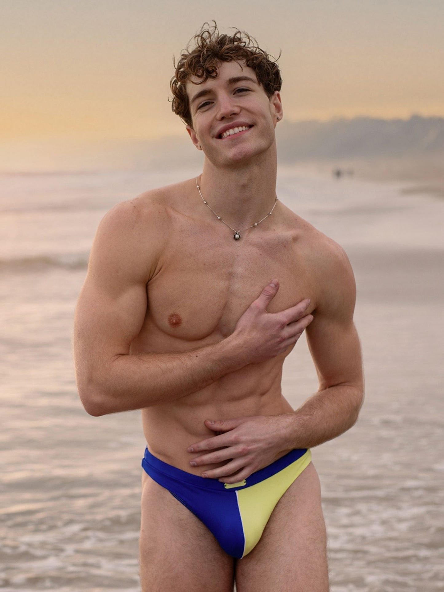 Man wearing blue and yellow swim thong on a beach at sunset