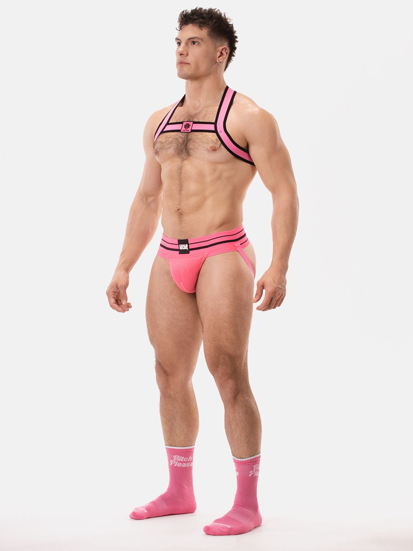 Man wearing a neon pink harness and jockstrap, with pink socks, on a white background