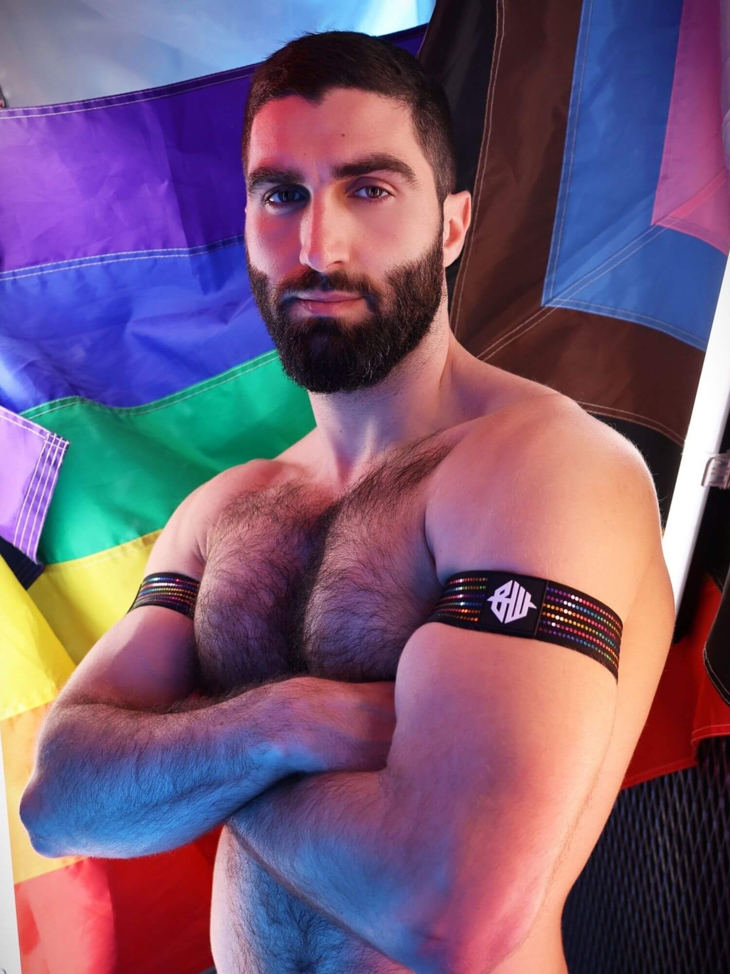Man with a beard and arm bands standing in front of a colorful flag