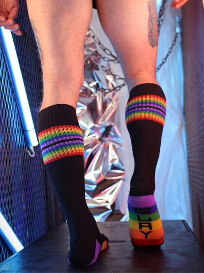 Person wearing black knee-high socks with rainbow stripes and colorful soles in a reflective, urban setting.