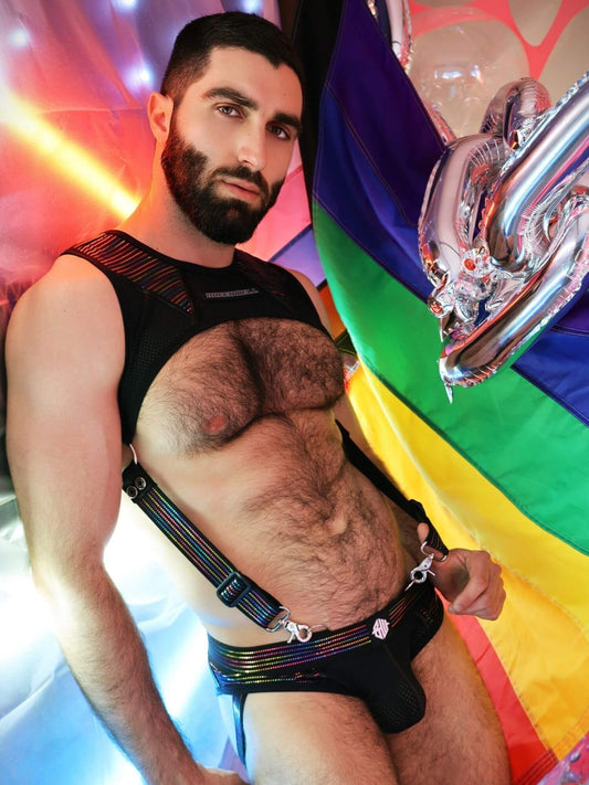 Man wearing a black tank top harness and colorful underwear against a rainbow flag background