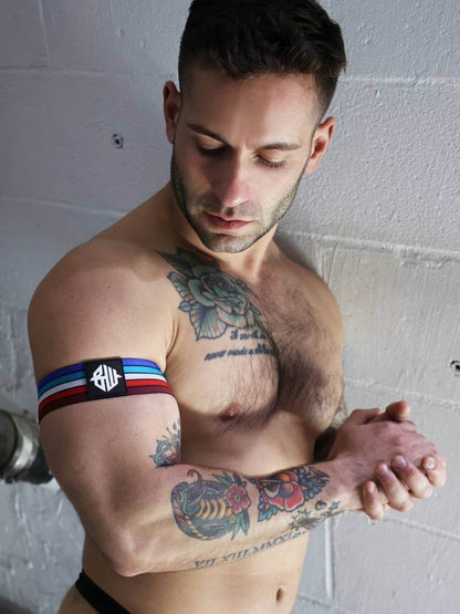 Man with tattoos wearing a black armband with red, white, and blue stripes against a white wall.