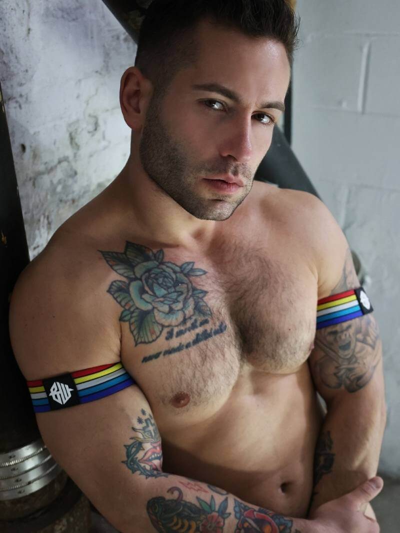 Man with tattoos and a rainbow wristband against a white wall.