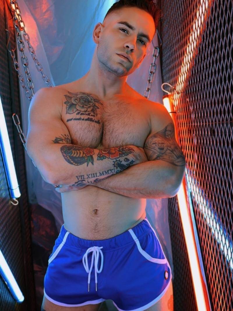 Man with tattoos wearing blue shorts in a dimly lit room with neon lights.