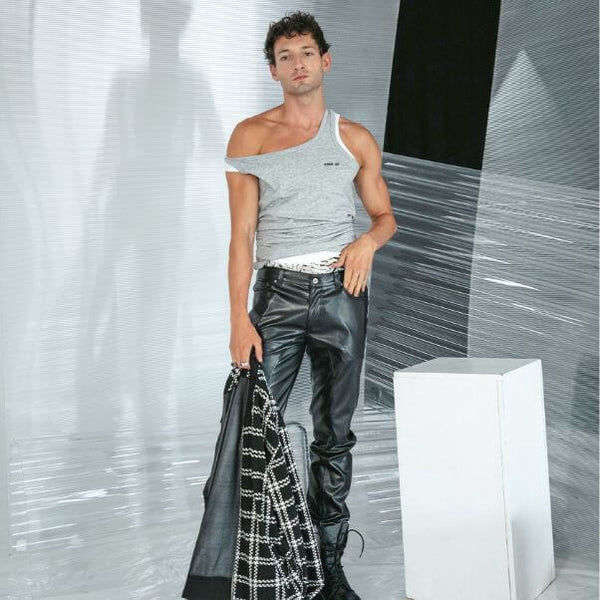 Man wearing a gray tank top and black pants holding a jacket against a textured wall.