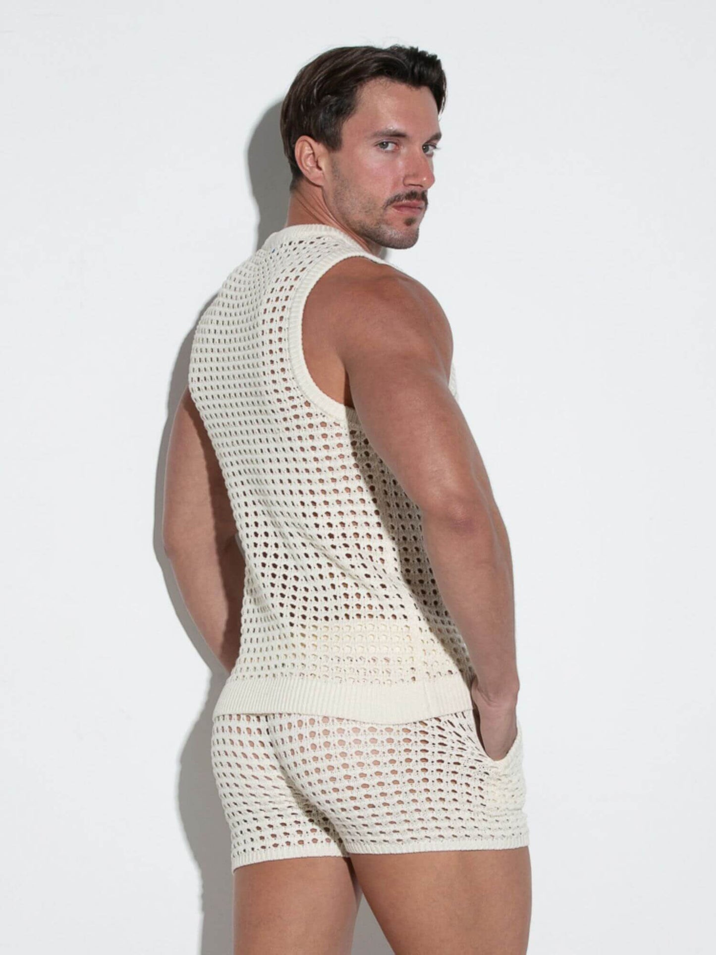 Man wearing a white crochet tank top and shorts against a light background