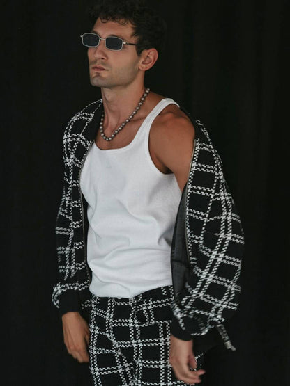 Man wearing a white tank top, black and white patterned cardigan, and checkered pants against a black background.