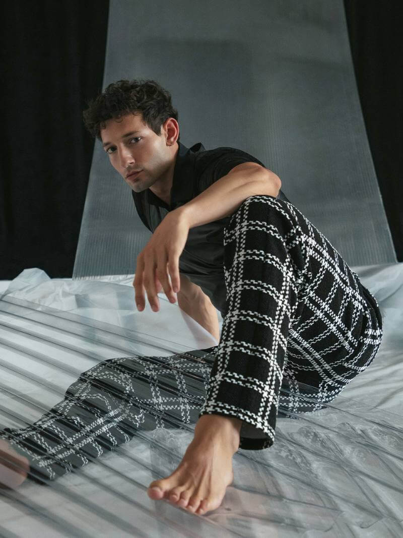 Man sitting on a bed wearing black and white checkered pants against a neutral background