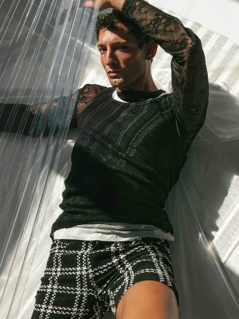 Person wearing a black lace top and plaid shorts with tattoos, standing against a white curtain.