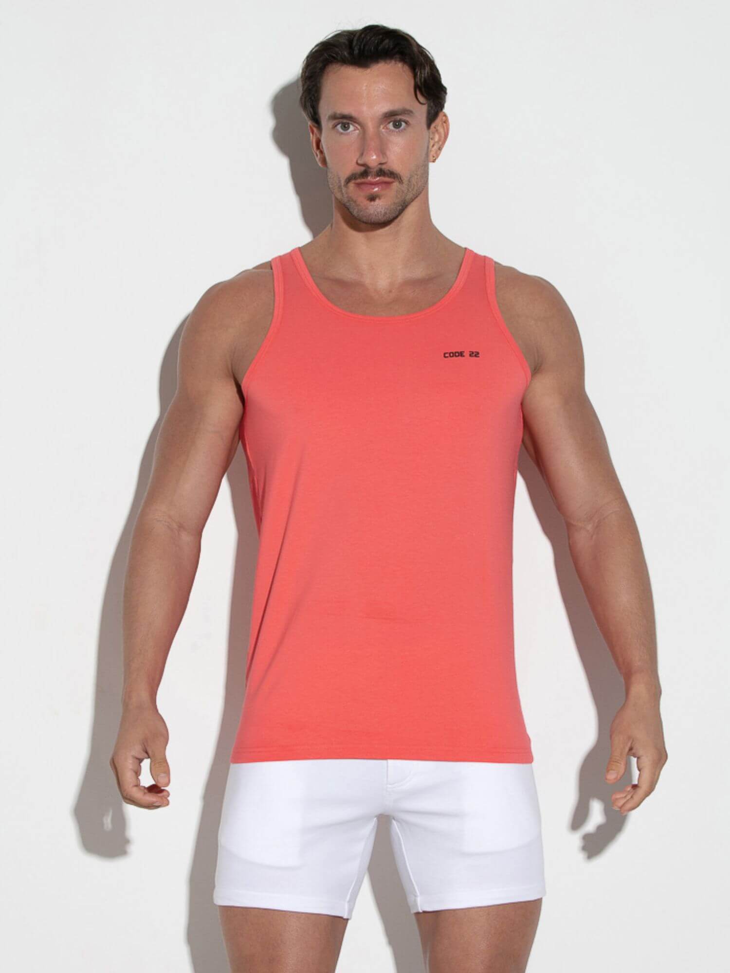 Man wearing a coral tank top and white shorts on a light gray background