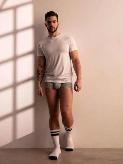 Man wearing a white t-shirt and underwear with socks standing against a beige wall.