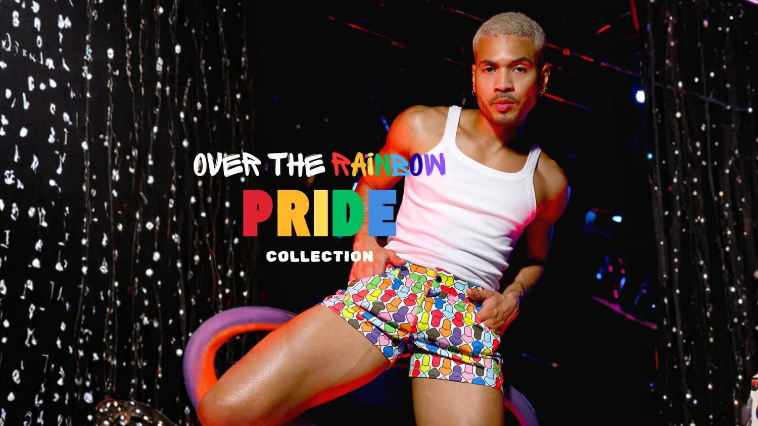 Person wearing a white tank top and colorful MODUS VIVENDI Rainbow Hearts shorts with 'Over the Rainbow Pride Collection' VOCLA pride menswear collection text.