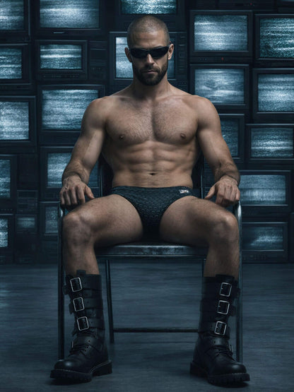 Man in black underwear and boots sitting on a chair with a dark, futuristic background.
