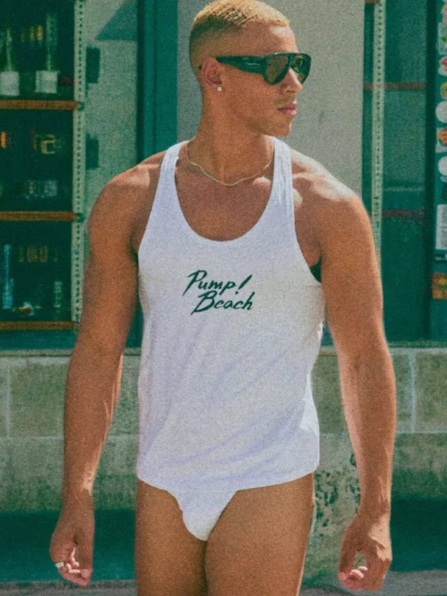 Man wearing a white tank top with 'Pump Beach' text in a casual setting.