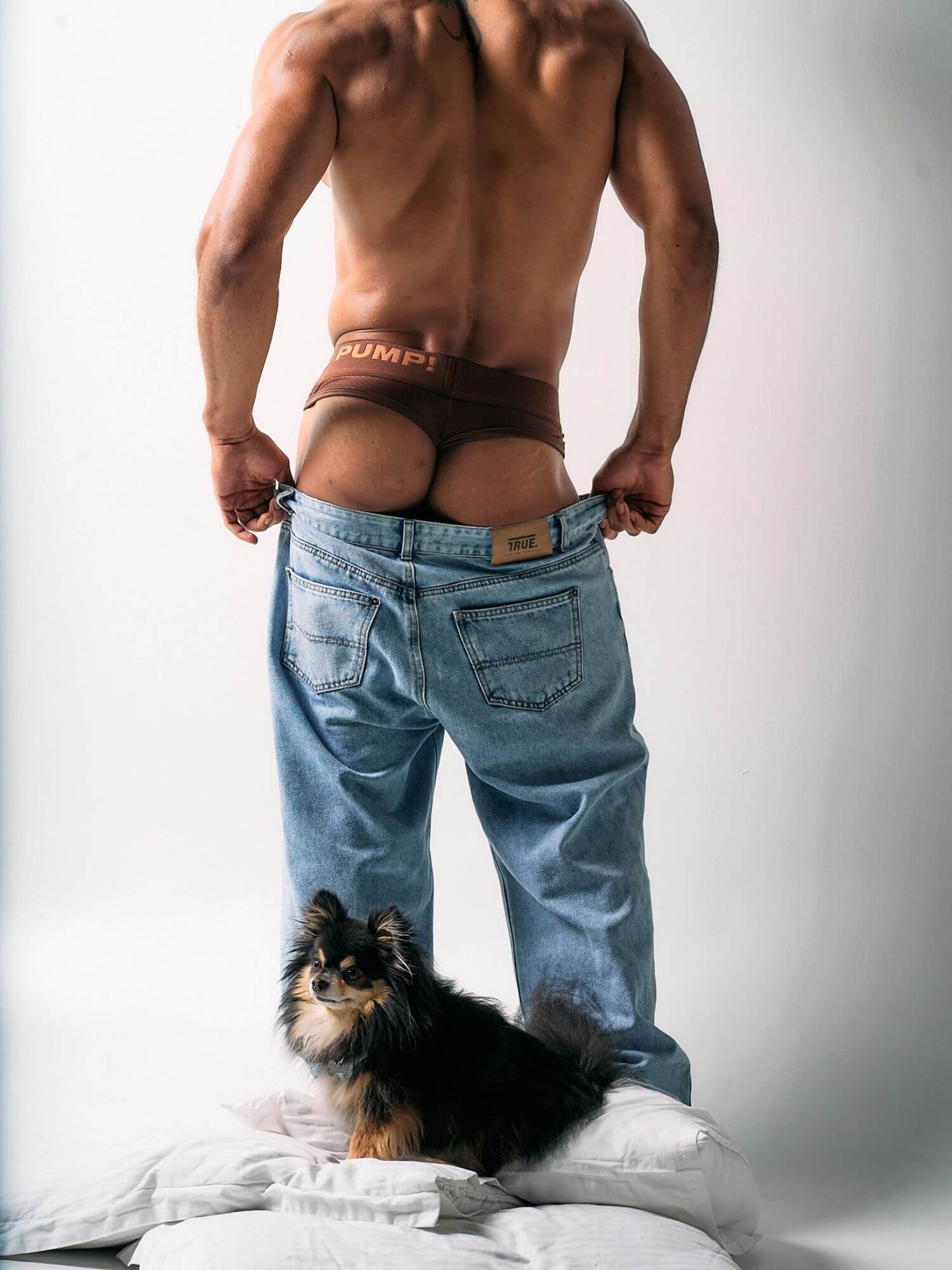 Man wearing a red thong and blue jeans with a small dog on a bed.