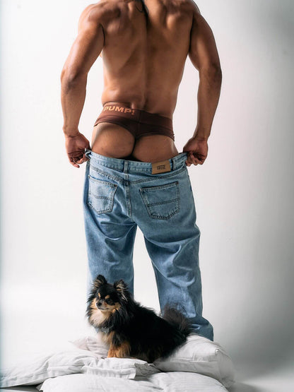 Man wearing a red thong and blue jeans with a small dog on a bed.