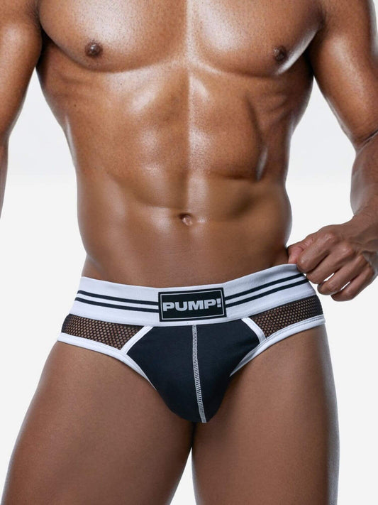 A close-up of a muscular male model wearing low-rise swim briefs. The briefs are black with white trim, featuring a front mesh panel and a white waistband with black stripes and a central black label that reads 'PUMPI'.