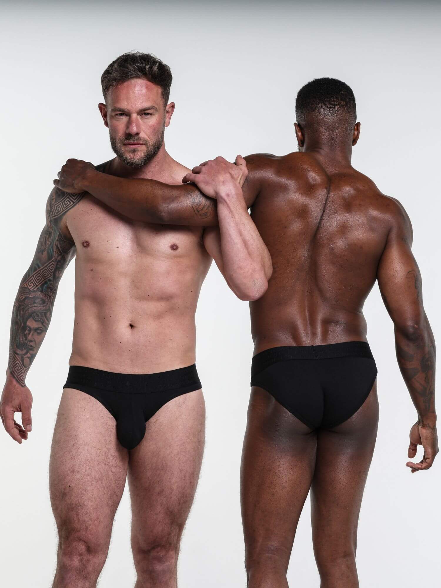 Two men wearing black underwear standing back-to-back against a white background