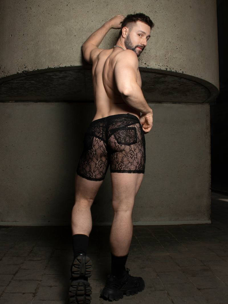 Man wearing black lace underwear standing against a textured wall.